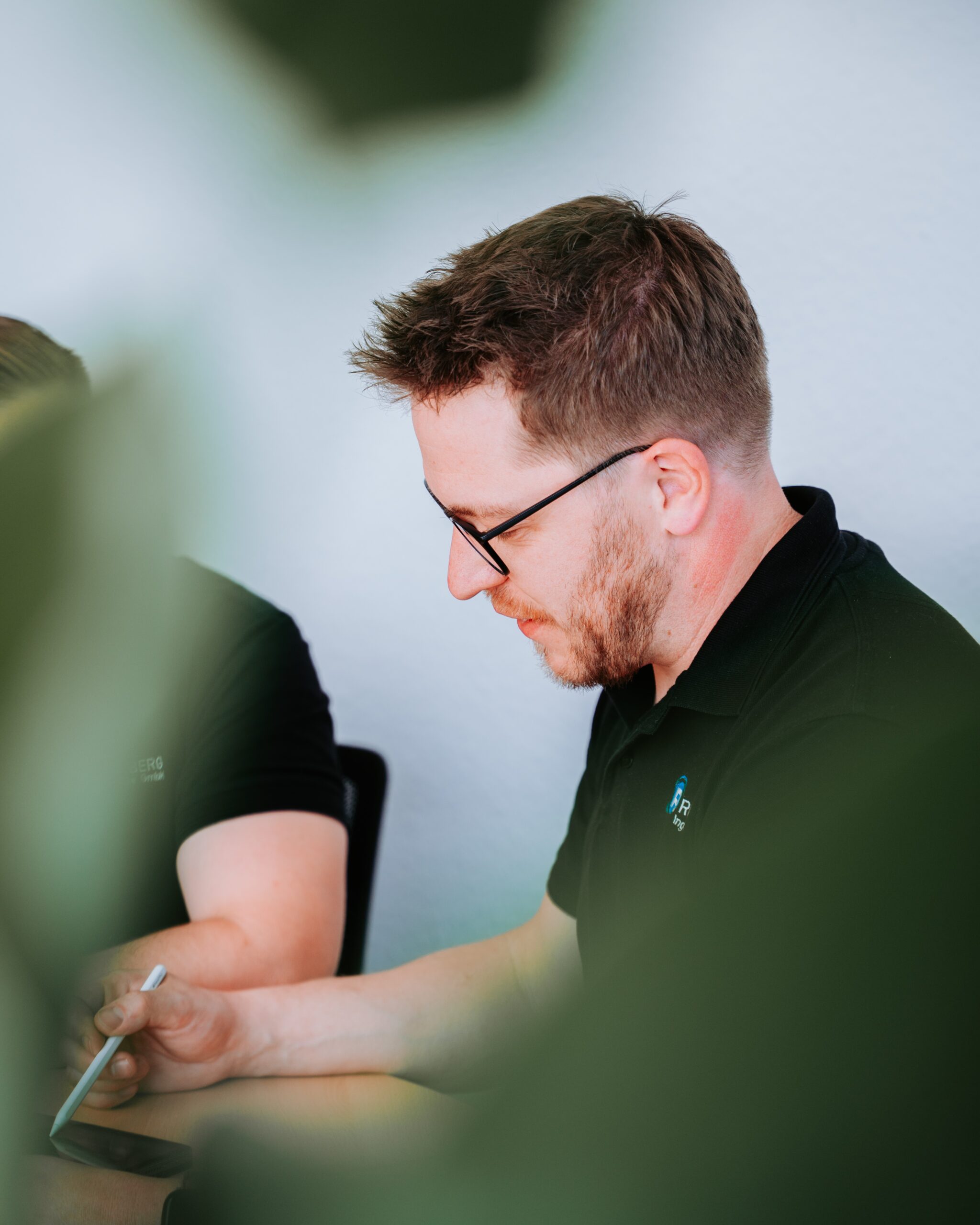 Zu sehen ist eine Person die mit einem Stift auf einem Bildschirm skizziert. Die Person trägt dunkle Kleidung und eine Brille. Unscharf im Vordergrund ist eine Pflanze erkennbar.