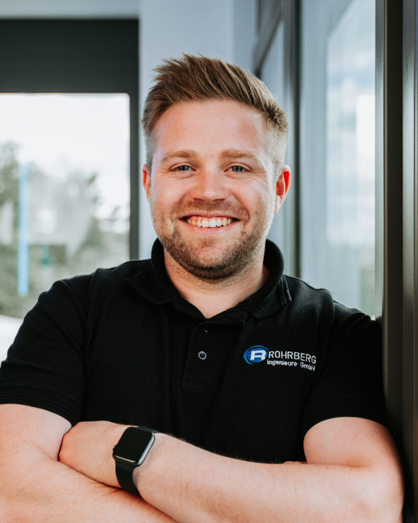 Ein Portrait einer der Mitarbeiter von Rohrberg Ingenieure. Die Person lächelt in die Kamera und trägt dunkle Kleidung und hat die Arme verschränkt.