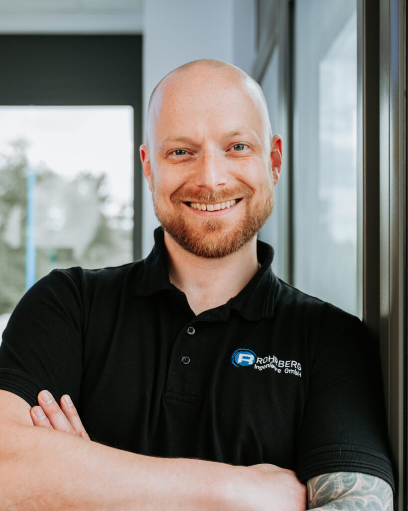 Ein Portrait einer der Mitarbeiter von Rohrberg Ingenieure. Die Person lächelt in die Kamera und trägt dunkle Kleidung und hat die Arme verschränkt.