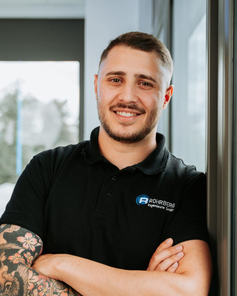 Ein Portrait einer der Mitarbeiter von Rohrberg Ingenieure. Die Person lächelt in die Kamera und trägt dunkle Kleidung und hat die Arme verschränkt.