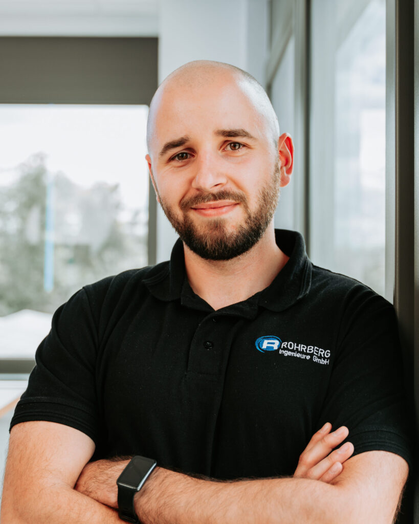 Ein Portrait einer der Mitarbeiter von Rohrberg Ingenieure. Die Person lächelt in die Kamera und trägt dunkle Kleidung und hat die Arme verschränkt.