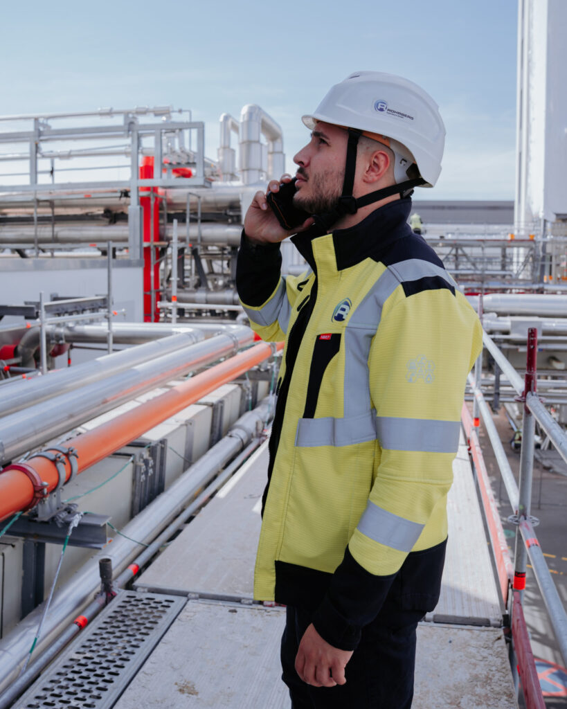 Ein Mitarbeiter von Rohrberg Ingenieure steht auf einem Gerüst und telefoniert. Die Person trägt Berufskleidung und einen Schutzhelm. Im Hintergrund sind Rohre und industrielle Anlagen zu sehen.