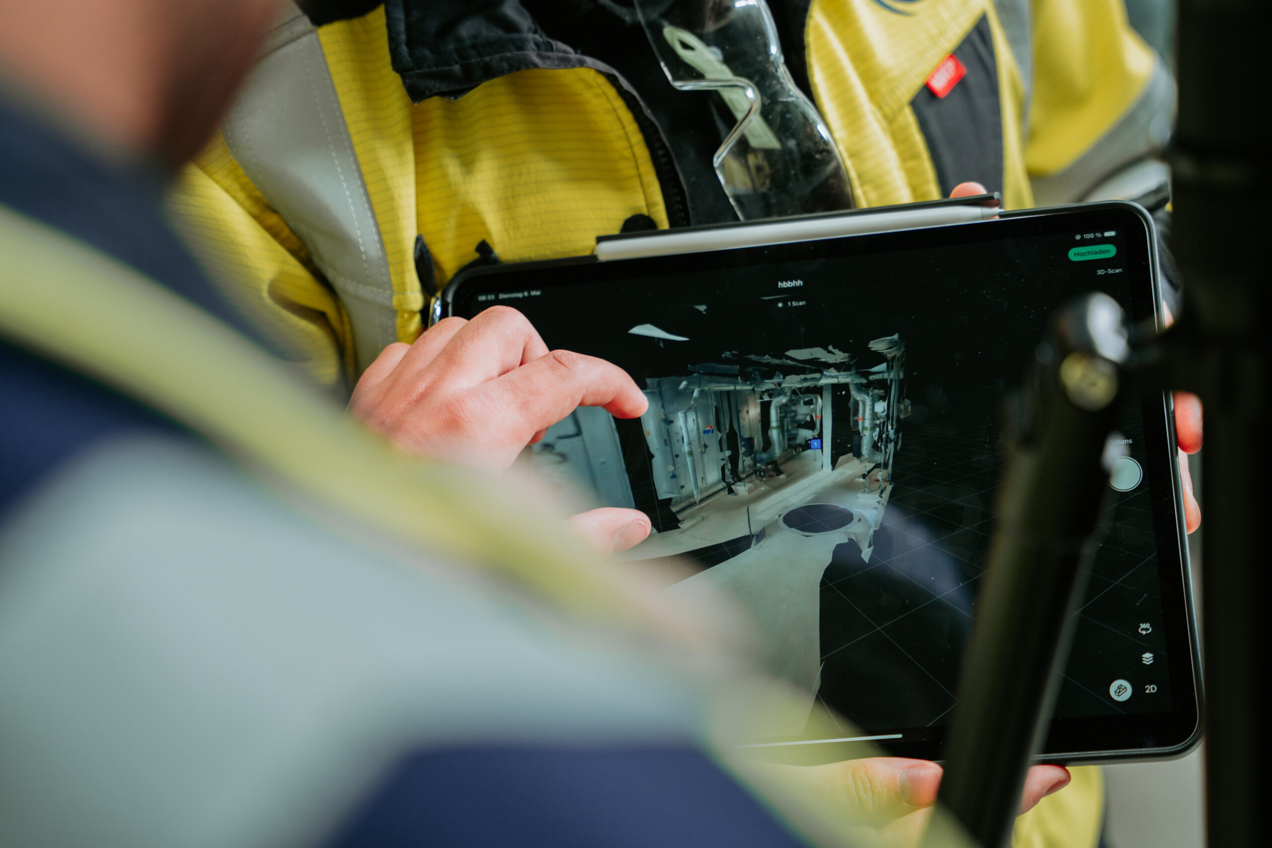 Zu sehen ist ein Tablet im Fokus, welches von mehreren, unscharfen Personen bedient wird. Auf dem Bildschirm ist ein Raumplan in 3D zu sehen.